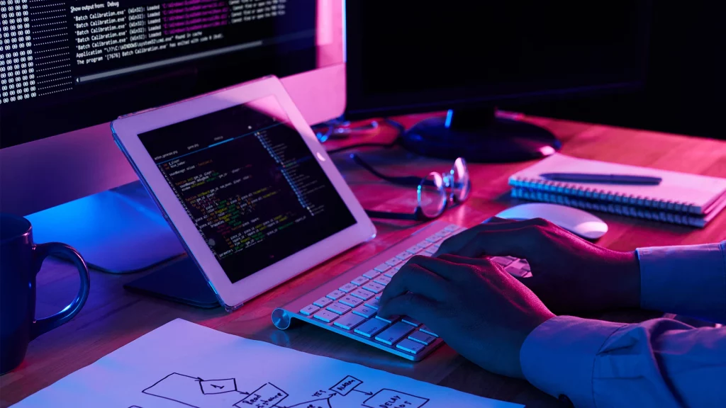 Una persona que trabaja en un escritorio con varias pantallas que muestran código de diseño web y una tableta, iluminada por el brillo de los monitores en una habitación oscura.