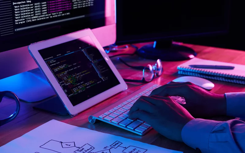 Una persona que trabaja en un escritorio con varias pantallas que muestran código de diseño web y una tableta, iluminada por el brillo de los monitores en una habitación oscura.